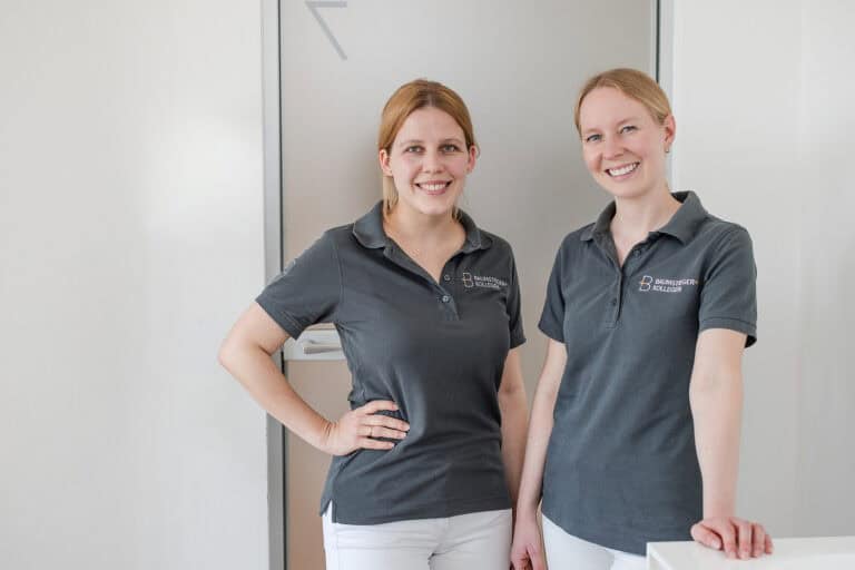Zwei lächelnde Kieferorthopädinnen in dunklen Polohemden mit der Aufschrift Baumstieger + Kollegen stehen angelehnt am Empfangstresen der Zahnarztpraxis.