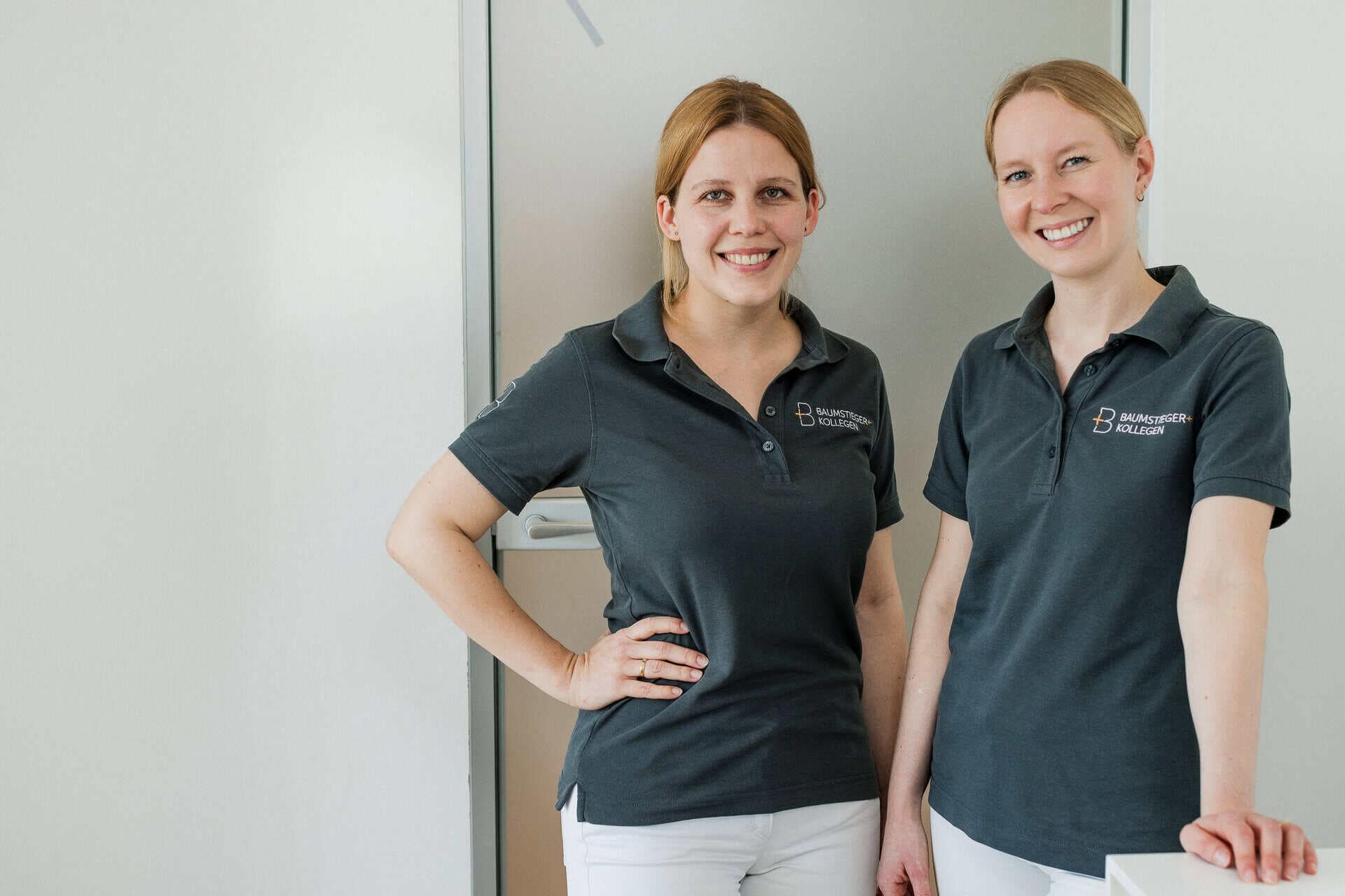 Zwei lächelnde Kieferorthopädinnen in dunklen Polohemden mit der Aufschrift Baumstieger + Kollegen stehen angelehnt am Empfangstresen der Zahnarztpraxis.