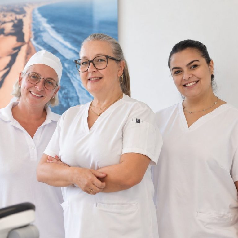 Lächelndes Prophylaxe-Team in weißen Kitteln bestehend aus drei Mitarbeiterinnen in Zahnarztpraxis mit freundlicher Atmosphäre.